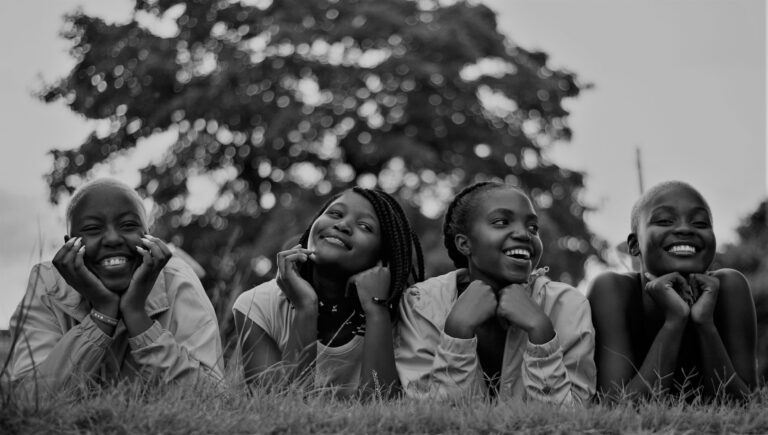 A group of four happy black women lying on grass, enjoying a joyful moment outdoors.